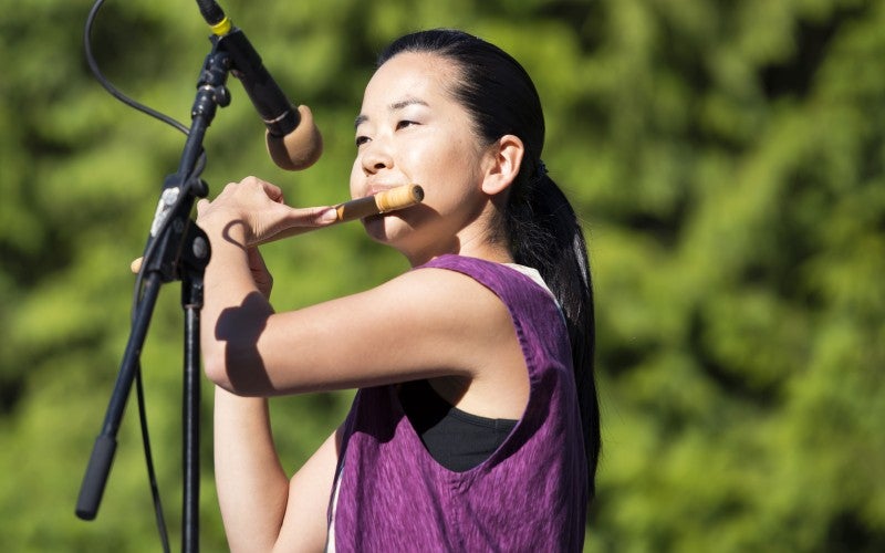 Kelsey stands in front of a microphone wearing a purple shirt and plays the fue (a wooden transverse flute).