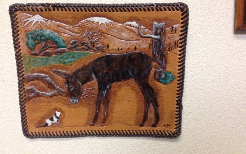 A hanging leather art piece depicting a brown donkey on a ranch, with mountains in the background.