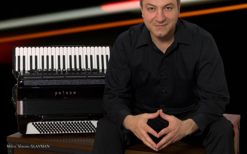 Milen Slavov wears a black shirt and sits next to a black accordion against a blurred background.