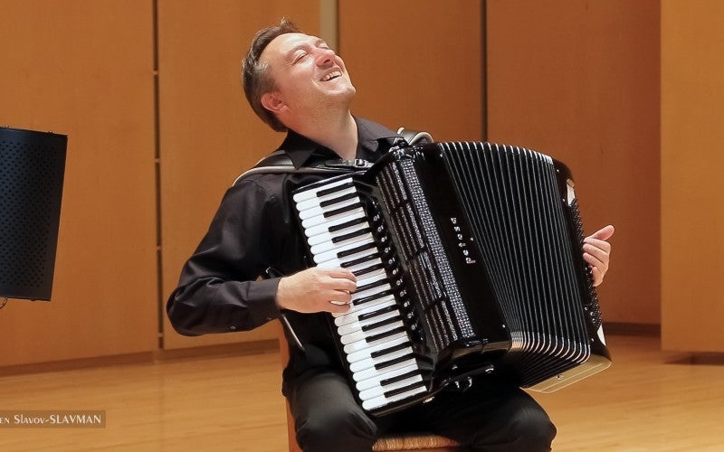 Milen Slavov wears a black dress shirt and pants and sits in a wooden chair playing a black accordion.