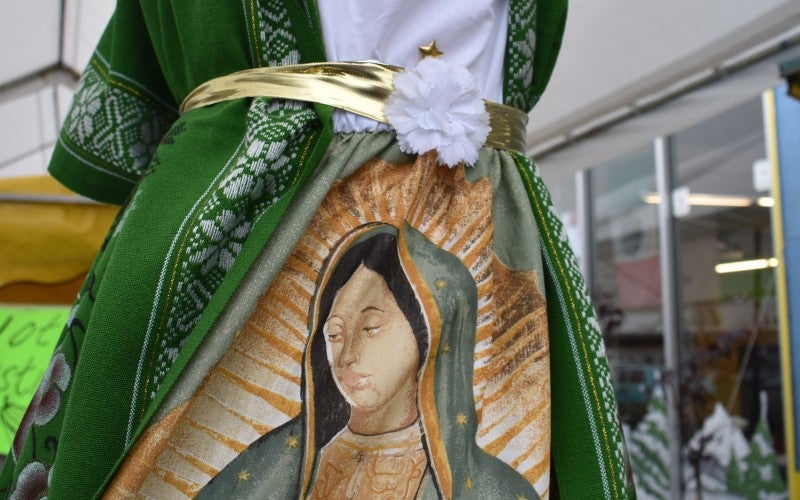 A white dress displayed on a mannequin featuring a long green cardigan, golden belt, and orange skirt with religious imagery.