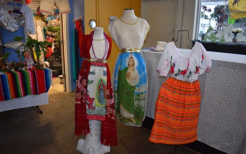 Three mannequins display traditional Mexican dresses. Each dress has a white upper portion and different colored skirts, including red, blue, and orange.