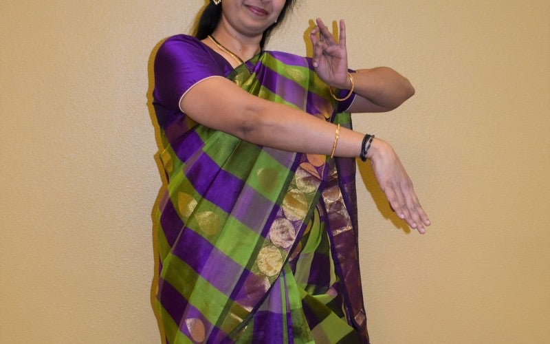 Anuradha demonstrates a traditional dance pose while wearing a purple and green traditional outfit.