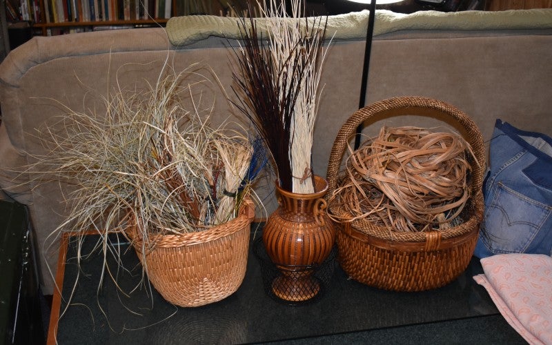 Two woven baskets, one tan and the other brown, filled with weaving material. A brown vase sits between them and holds more weaving material.