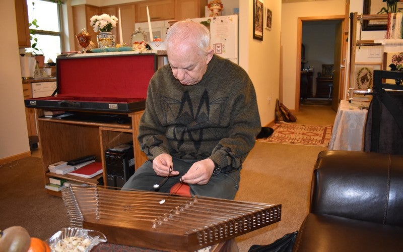 Jamshid sits while playing a wooden santur instrument and wears a green sweater.
