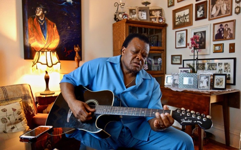 Norman Sylvester, a man, sits in a living room wearing a blue shirt and playing a black guitar.