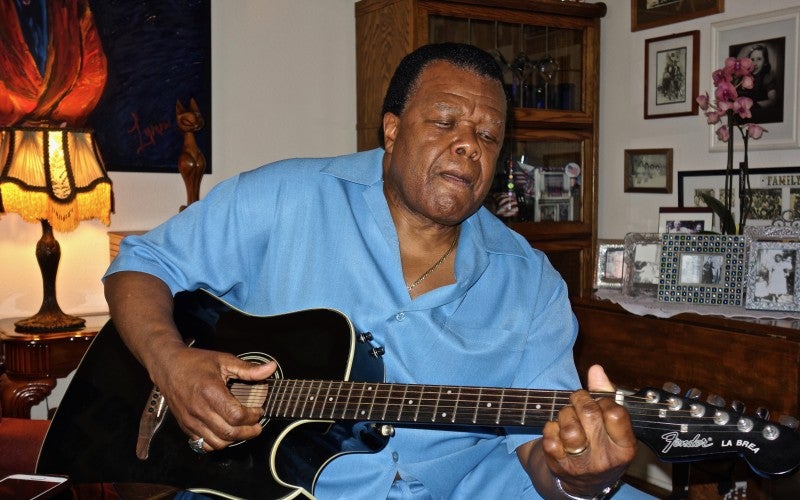 Norman Sylvester sits in a living room wearing a blue shirt and playing a black guitar.