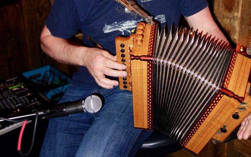 Johnny sits and plays an accordion. He wears a dark blue shirt and blue jeans.