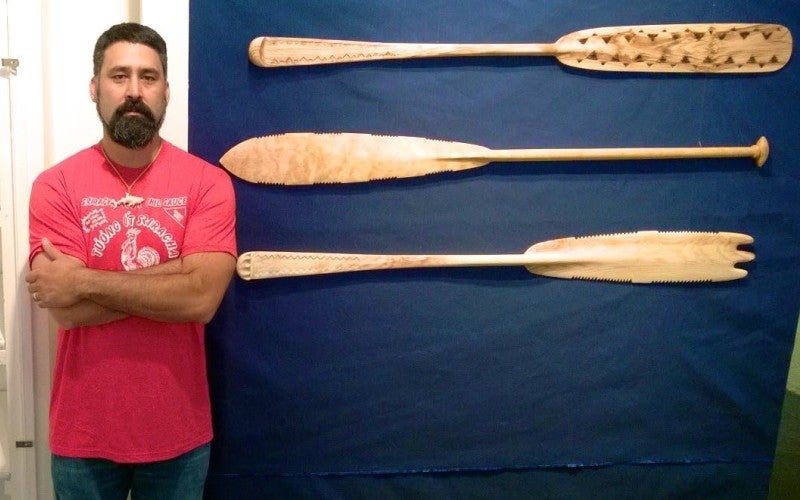 A man stands between 3 short wooden oars displayed on shelves and 3 long wooden oars displayed on a blue wall