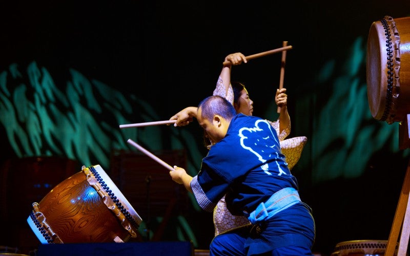 Two people play taiko drums on a stage. The person in front is wearing a blue traditional robe.