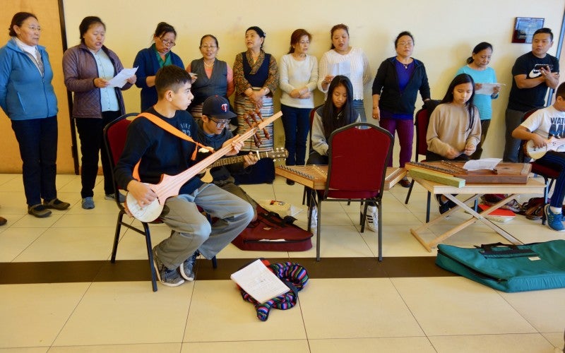 A person sits in a chair and plays a traditional stringed instrument, while a group of people stand behind them