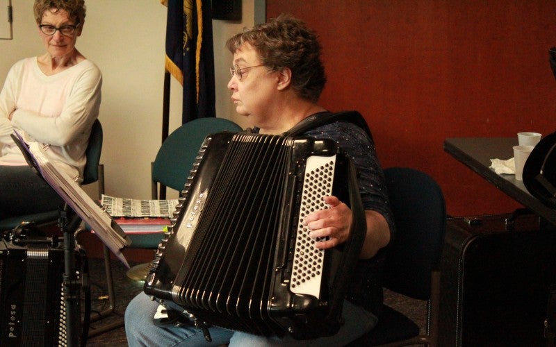 A woman sits in a blue chair and plays a black accordion.