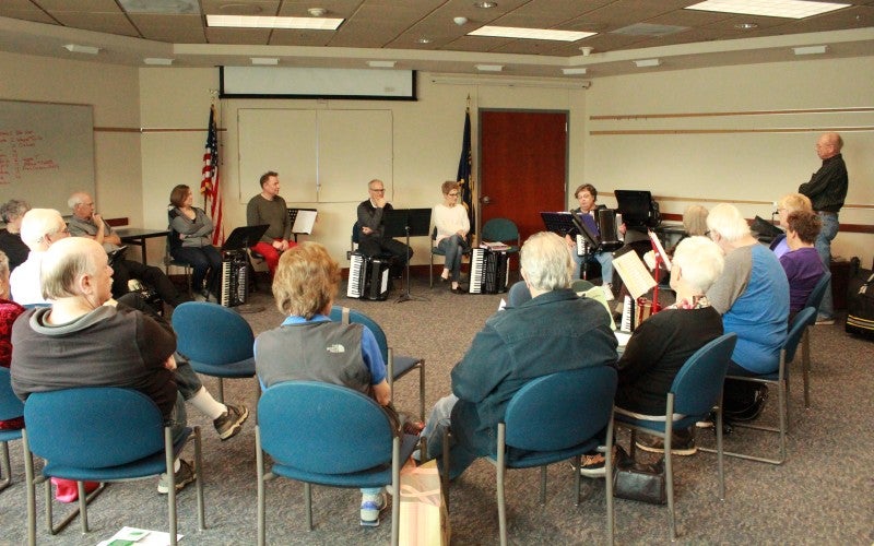 A group of people sit in a circle of chairs with music stands in front of them.