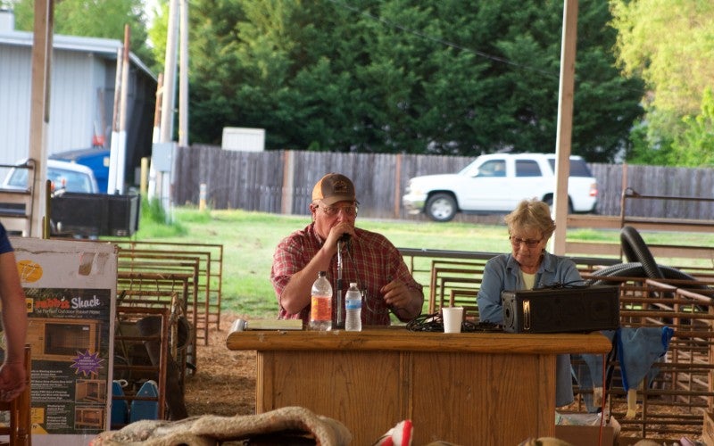 Jake sits at a wood table with a microphone during an outdoor live auction.
