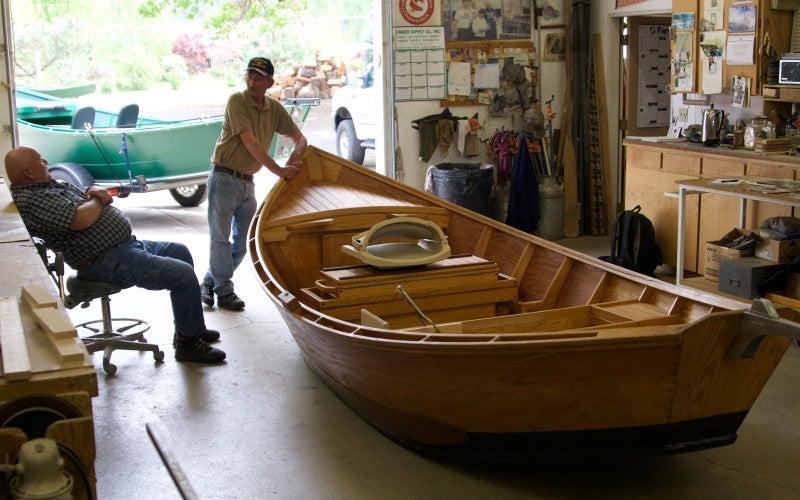 Two people stand in a garage next to a small wooden boat