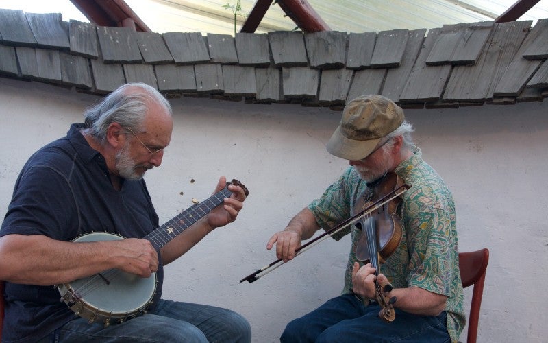 John and Joe sit outside playing a banjo and a fiddle.