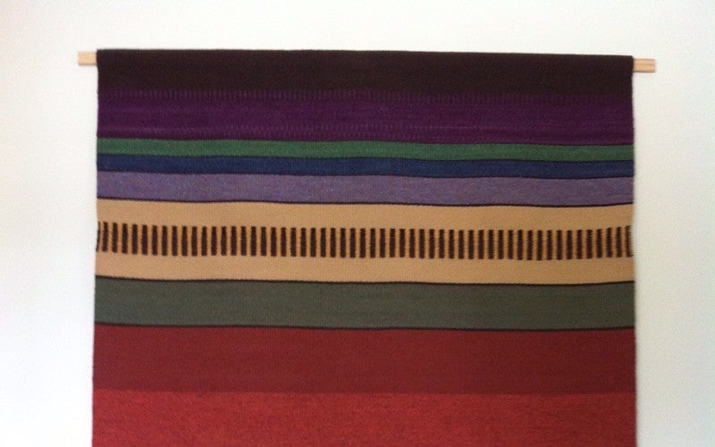Woven hanging blanket with horizontal bands of different colors like blue, purple, red, orange, yellow, green, and black.