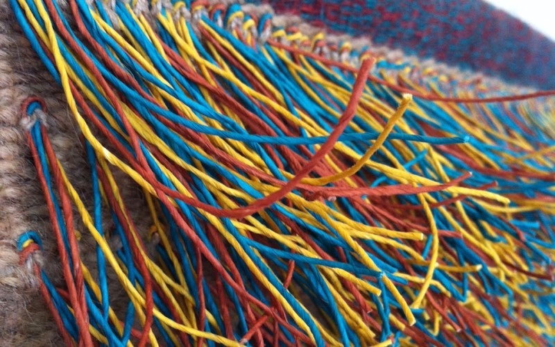 Close-up image of a multicolored fringe decoration on the end of a blue and red woven piece. The fringe is made up of blue, yellow, red, orange, and green threads.