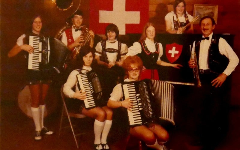 A group of seven people holding various instruments. A Switzerland flag hangs on the wall behind them.