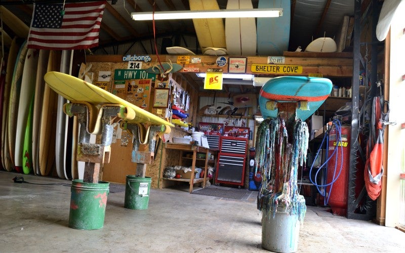 Brad's workshop, where a yellow and blue surfboard are on worktables, and the wall in the background is covered in various signs.