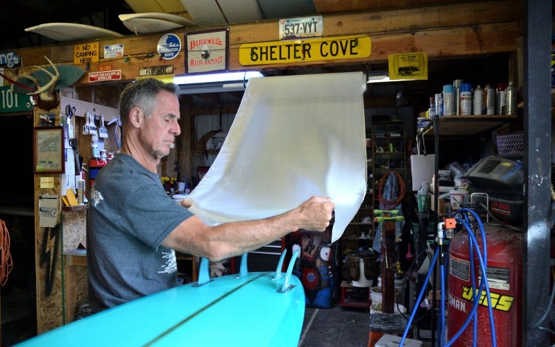 Brad stands working on a bright blue surfboard in his workshop.