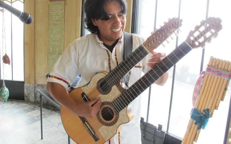  Alex Llumiquinga Perez holds a double necked guitar with a microphone and wooden pipes in the background.