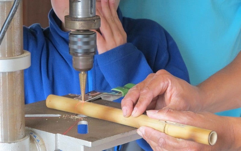 Person making a wooden pipe at a machine. A child watches wearing goggles and a blue fleece sweater.