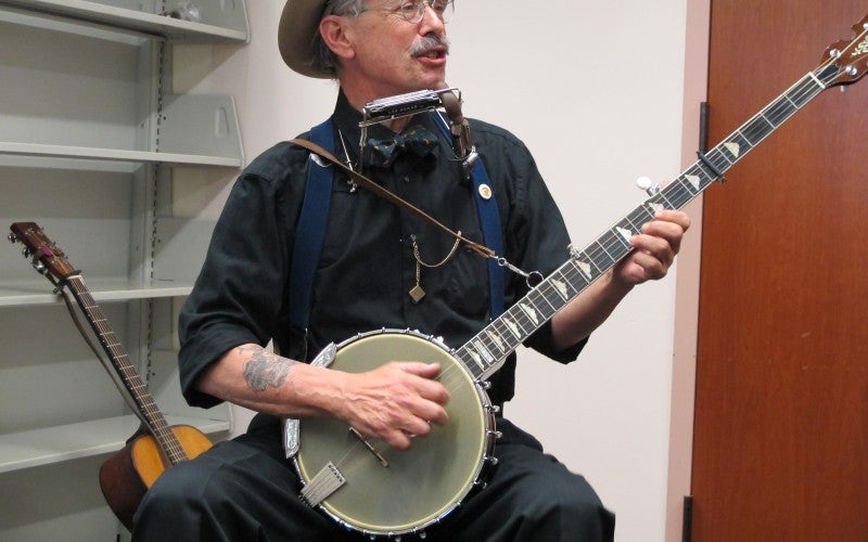 A man sits playing a banjo and wearing a black shirt and tan hat.