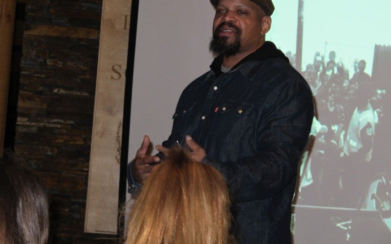 Crenshaw stands in front of a projector screen and gestures forward with his hands. He wears a gray collared shirt with a black hooded sweatshirt underneath, and a brown cap.