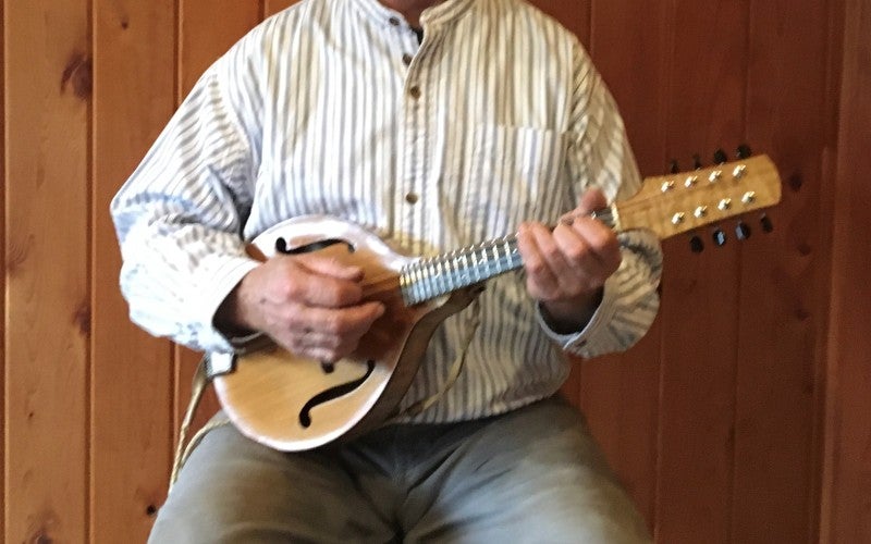 Bryan Ibach sits in front of a wood wall while holding a tan mandolin instrument and wearing a black hat, white striped shirt, and gray pants.