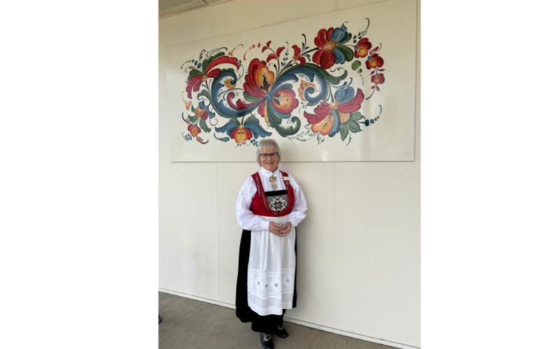 Meshnik stands in front of a rosemåling style mural. She wears a traditional Norwegian outfit.
