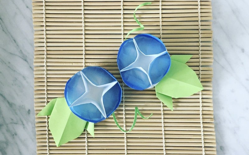 Two blue origami morning glory flowers rest on a bamboo mat