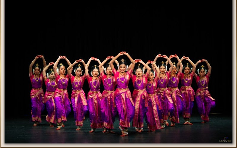 13 dancers are posing in expressive dance gesture while performing on stage. All of them in red and purple traditional Kuchipudi dance attire.