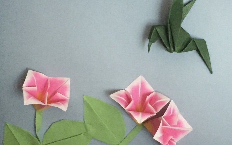 Green origami hummingbird hovers over pink origami flowers