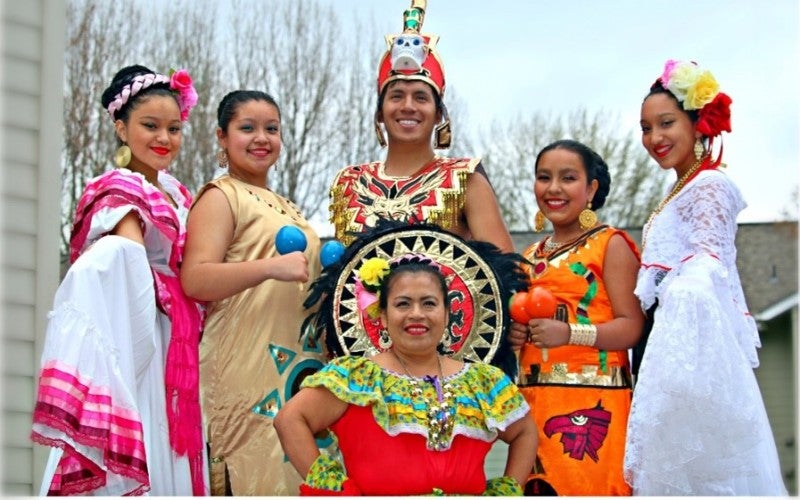 Luisa Valentín Pelagio posing with her group of dancers all dressed in folklorico dance costumes. They are outside with trees in the background.