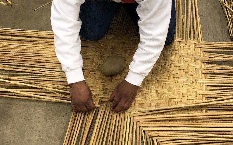 Dagoberto kneeling on the floor weaving a Tule mat.