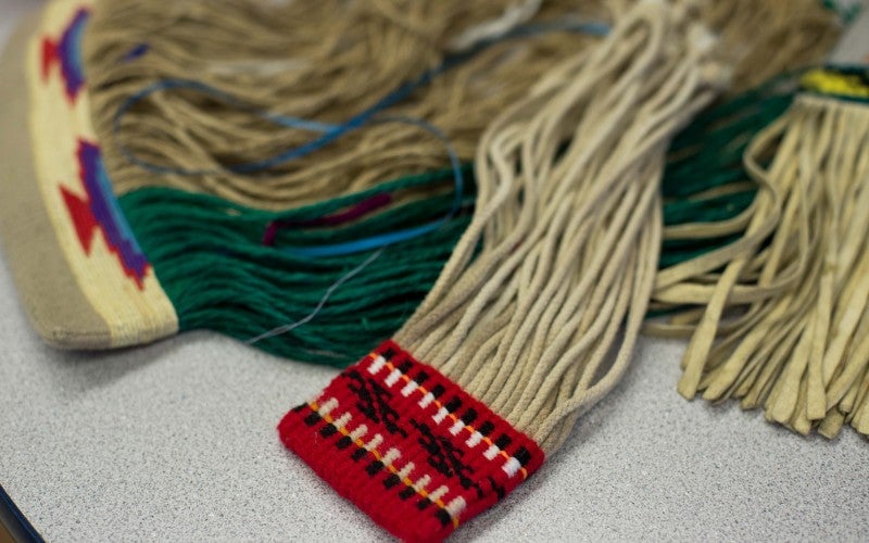 Several woven items sitting on a table. One is red with black and white patterning and strings coming out of it and another has wood with red, black and blue patterning and green and tan strings coming out of it.