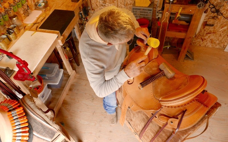 Bill stands in his workshop crafting a tan Western-style saddle, and wears a gray long sleeve shirt and blue jeans.
