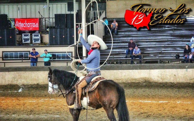 Josue Mendoza riding a horse and performing rope skills. He is wearing a blue shirt, white hat, and leather chaps.