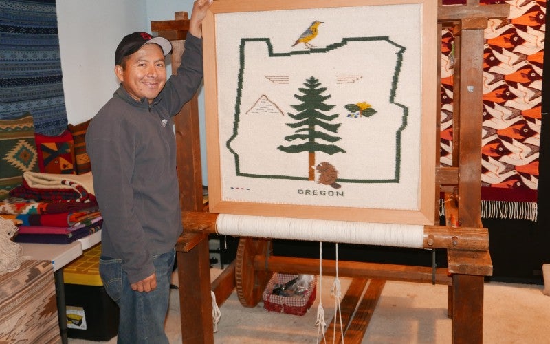 Francisco stands next to a large white rug with a green outline of the state of Oregon. In this outline are Oregon related images: a beaver, a Douglas fir, a Western meadowlark bird, and an Oregon Grape plant.
