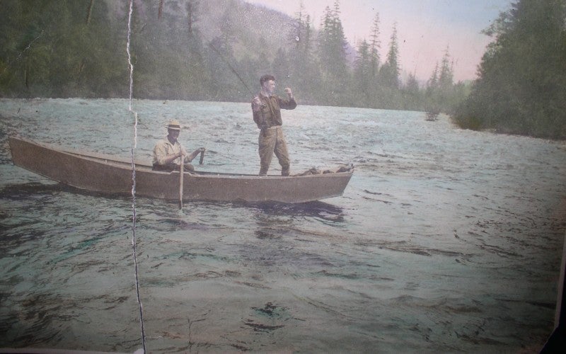 2 men on a fishing boat in the middle of the river. One man is standing and is about to cast a line, he is holding a fishing rod.