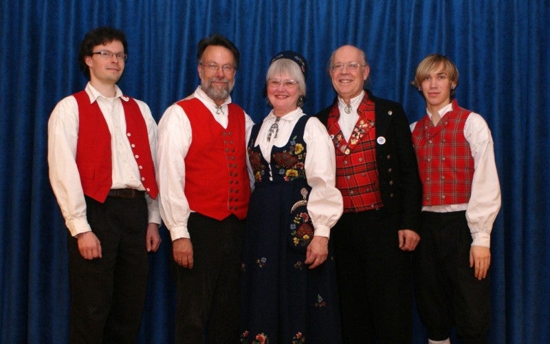 Fossegrimen poses against a blue curtain. They wear traditional vests over white shirts with black pants. Claire Elliker-Vågsberg wears a long black embroidered dress.