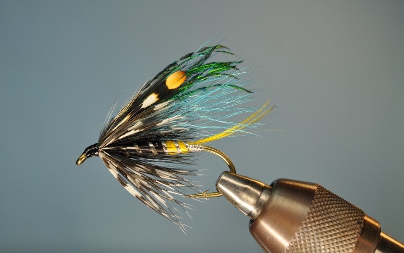 A black, yellow, and green fly fishing hook against a grey background.