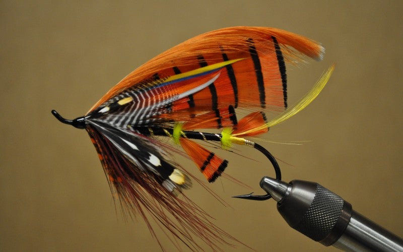 A black, yellow, and red fly fishing hook against a brown background.