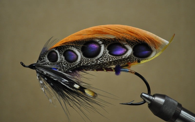 A black, yellow, purple, and orange fly fishing hook against a brown background.