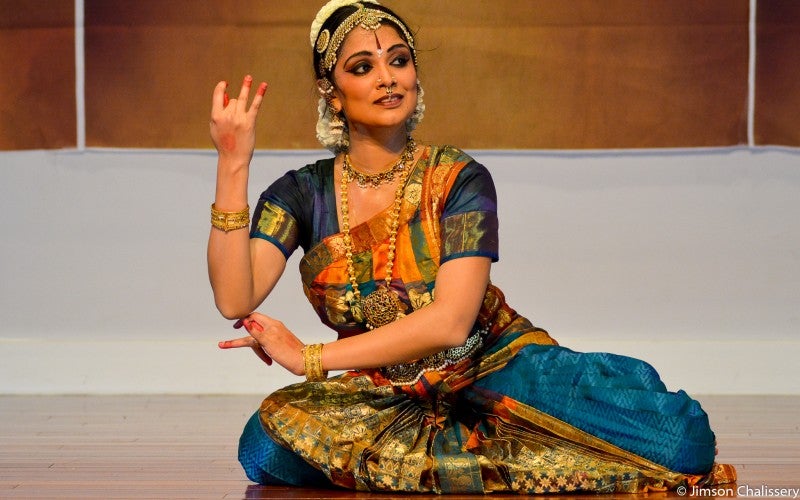 Sweta dancing, posed on her knees with her right hand raised and left arm crossing over her torso, looking up