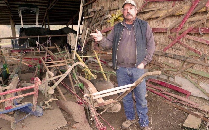 Dave stands next to multiple horse-drawn plows and wears a brown sweater, black vest, blue jeans, and tan baseball cap.