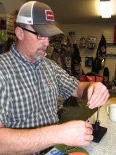 John Garrett stands in the Gorge Fly Shop in Hood River, Oregon, and ties a fly. He wears a gray plaid shirt and a brown baseball cap.