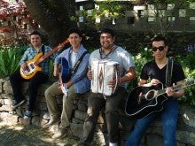 Four members of Los Amigos de la Sierra sit outside on a stone wall and each hold various instruments, including a bass guitar, two acoustic guitars, and an accordion.