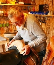 Bill Huston stands in his workshop and adjusts the back of a black saddle. He wears a light gray long sleeved shirt and a light yellow bandana around his neck.